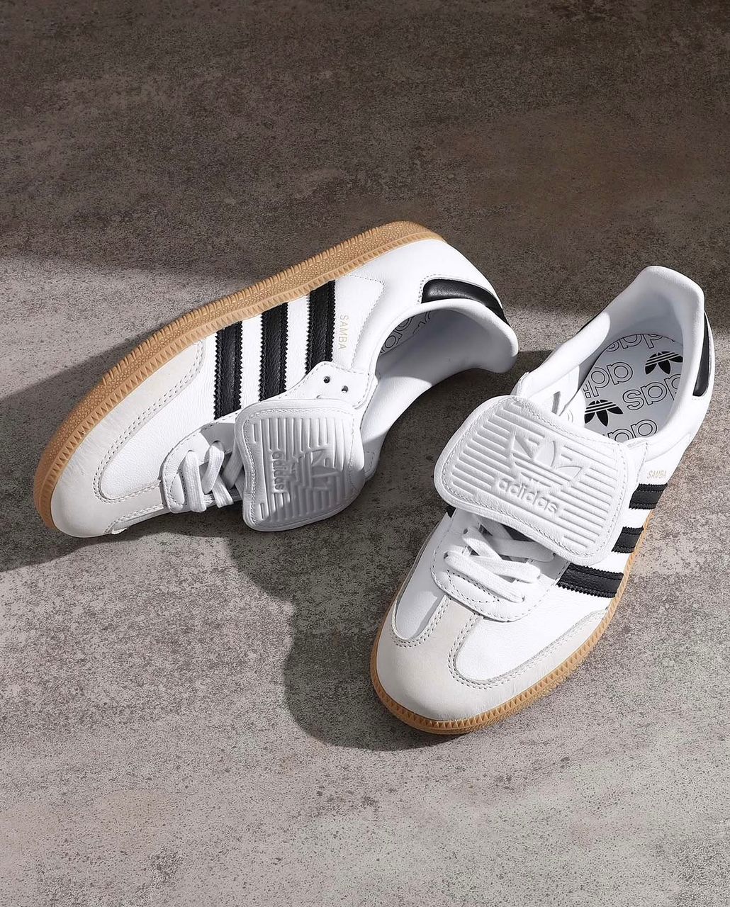 Adidas Samba OG sneakers Adidas Samba OG men's sneakers in white leather with black Three Stripes, gum rubber sole, and white velcro strap, displayed on concrete surface.