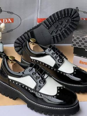 Men's black and white patent leather spectator derby shoes with silver stud detailing and thick black chunky lug sole, displayed with shoe trees and branded boxes.