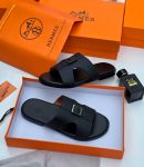 Hermès men’s black Epsom leather Oran slide sandals with signature H-cutout, displayed in orange Hermès box with branded dust bag and perfume bottle on white surface.