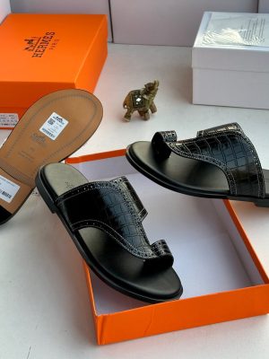 Hermès men's black crocodile-embossed leather Izmir thong sandals displayed with signature orange Hermès box and gold elephant figurine on white surface.