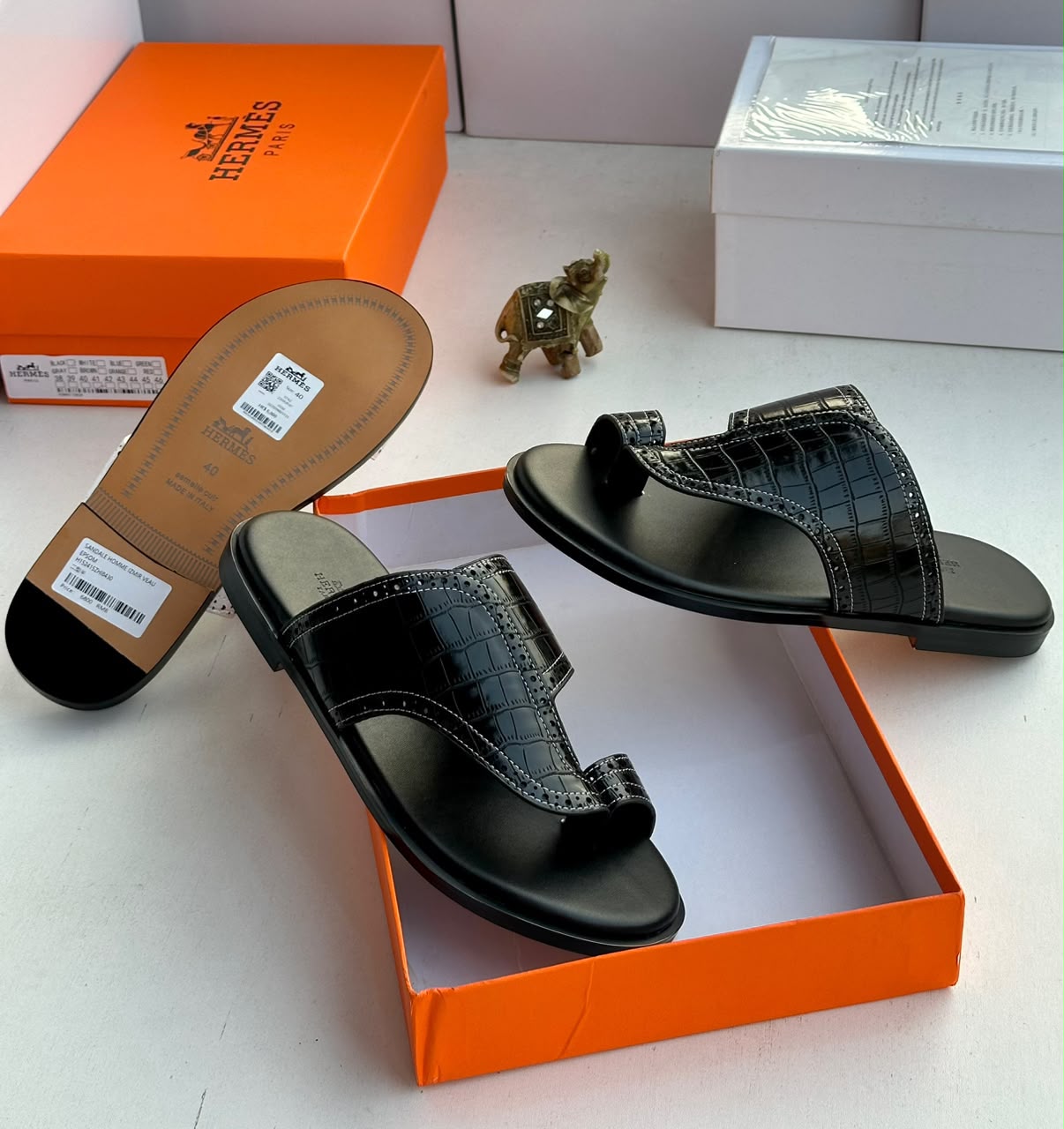 Bold French luxury Hermès sandals Hermès men's black crocodile-embossed leather Izmir thong sandals displayed with signature orange Hermès box and gold elephant figurine on white surface.