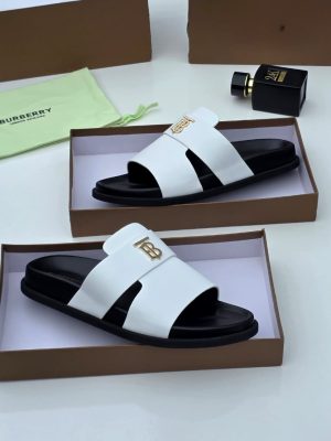 Burberry men's black leather slide sandals with gold TB monogram hardware, displayed in branded brown boxes with lime green dust bags and perfume bottle.