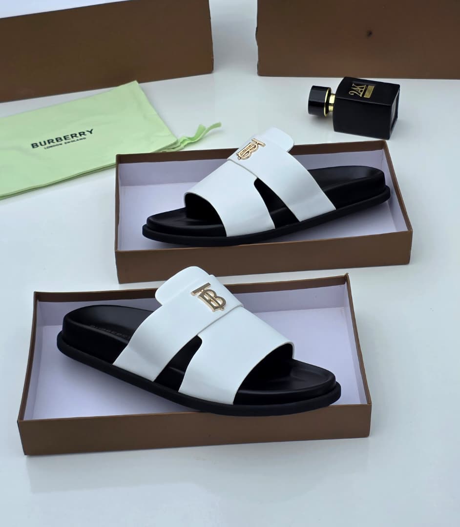 Premium Burberry black leather slides Burberry men's black leather slide sandals with gold TB monogram hardware, displayed in branded brown boxes with lime green dust bags and perfume bottle.
