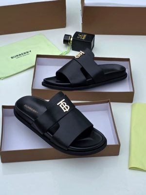 Burberry men's black leather slide sandals with gold TB monogram hardware, displayed in branded brown boxes with lime green dust bags and perfume bottle.