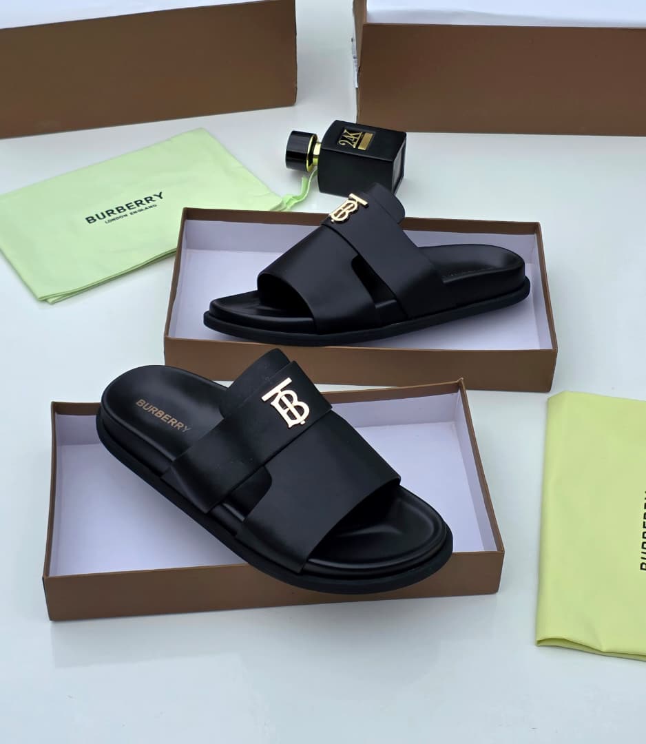 Premium Burberry black leather slides Burberry men's black leather slide sandals with gold TB monogram hardware, displayed in branded brown boxes with lime green dust bags and perfume bottle.