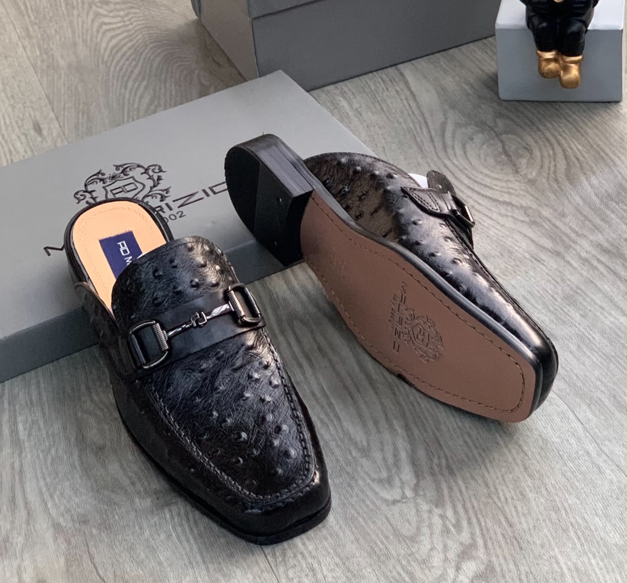 Genuine ostrich leather horsebit mules Men's brown ostrich leather horsebit mules with square toe and stacked heel, displayed on grey wood floor with branded shoebox.