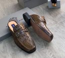 Men's brown ostrich leather horsebit mules with square toe and stacked heel, displayed on grey wood floor with branded shoebox.