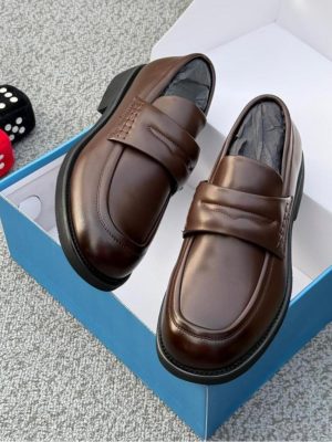 Men's chocolate brown leather penny loafers with black rubber sole, displayed in open blue shoebox with decorative dice on grey carpet.