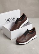 Hugo Boss men's brown pebbled leather slip-on sneakers with black elastic panels and white midsole, displayed with branded white shoebox on a grey surface.