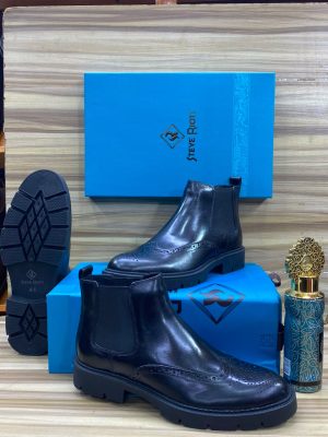 Men's black leather Steve Riots combat boots with thick ridged platform sole, displayed on wooden surface with branded blue shoebox and decorative perfume bottle.