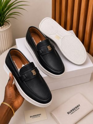 Burberry men's tan leather slip-on loafers with gold TB monogram hardware and white rubber sole, held in hand with branded white shoebox and dust bag on beige surface.