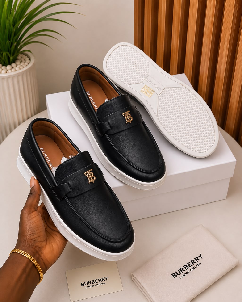 Burberry tan leather slip-on loafers Burberry men's tan leather slip-on loafers with gold TB monogram hardware and white rubber sole, held in hand with branded white shoebox and dust bag on beige surface.