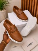 Burberry men's tan leather slip-on loafers with gold TB monogram hardware and white rubber sole, held in hand with branded white shoebox and dust bag on beige surface.