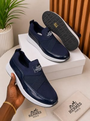 Hermès men's navy blue slip-on sneakers with patent leather accents, white rubber sole, and branded hardware, held in hand with white shoebox and dust bag on beige surface.