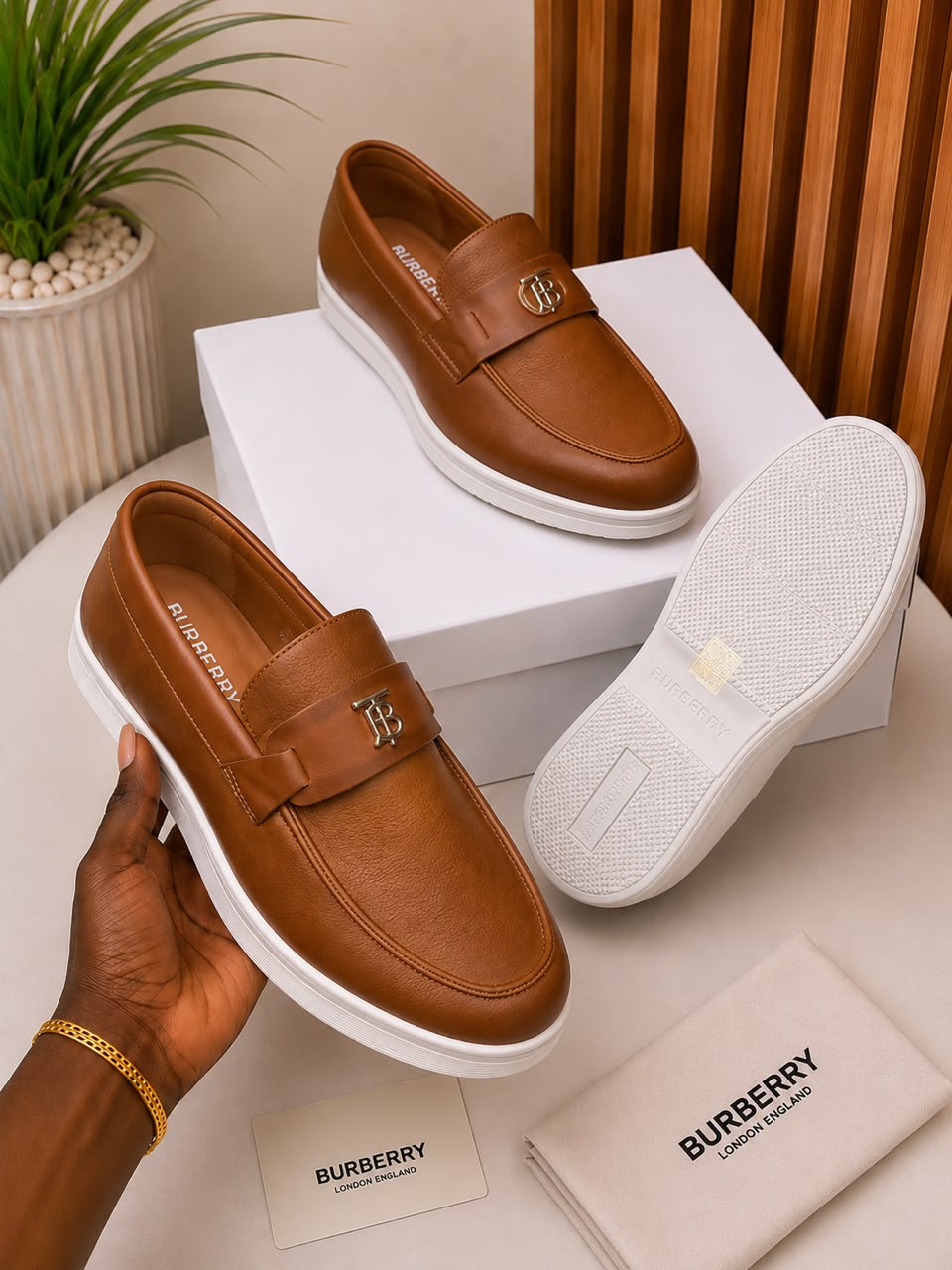 Burberry tan leather slip-on loafers Burberry men's tan leather slip-on loafers with gold TB monogram hardware and white rubber sole, held in hand with branded white shoebox and dust bag on beige surface.