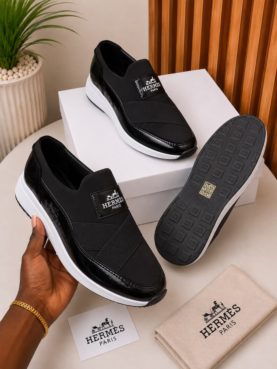 Hermès slip-on sneakers Hermès men's navy blue slip-on sneakers with patent leather accents, white rubber sole, and branded hardware, held in hand with white shoebox and dust bag on beige surface.