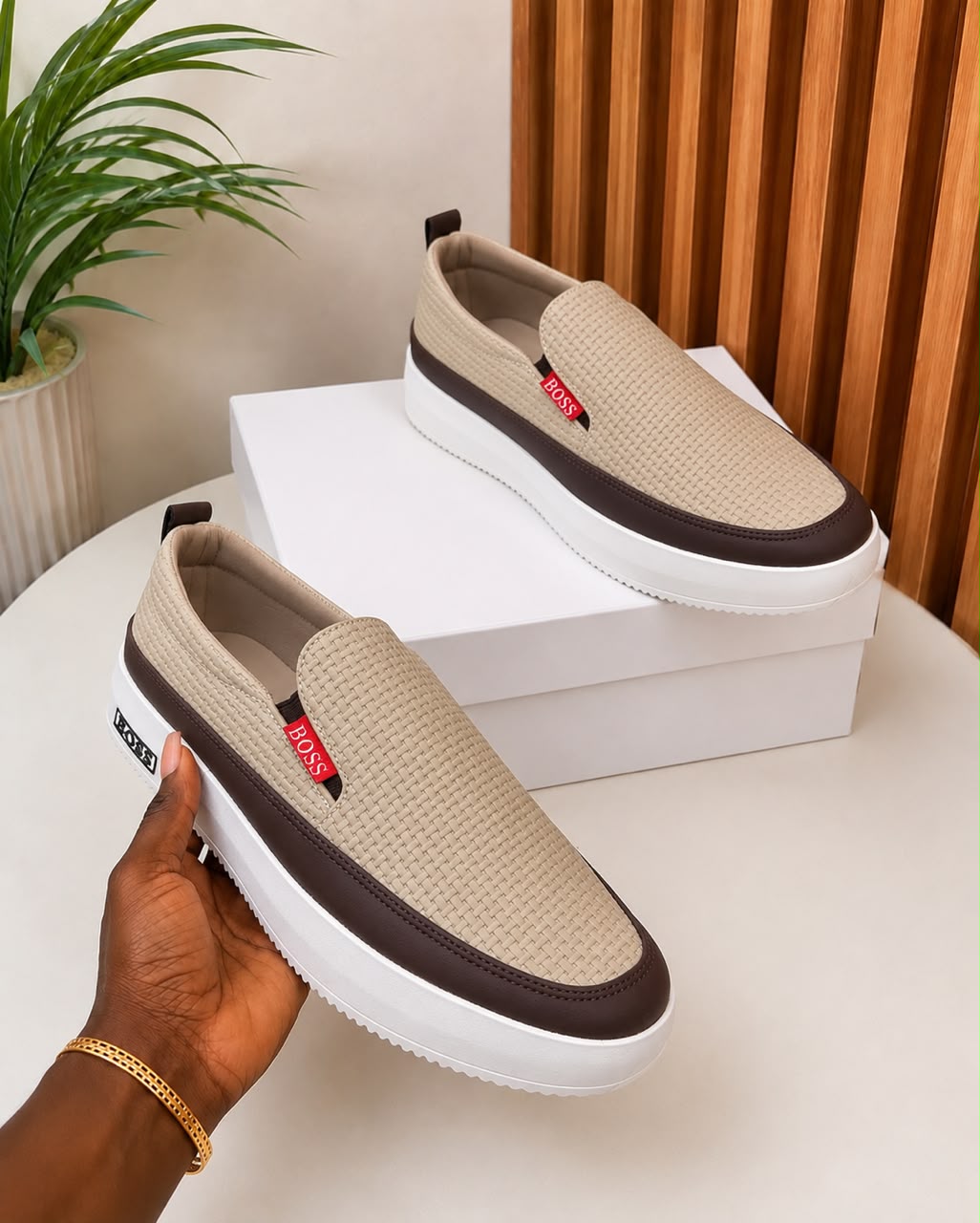 Hugo Boss woven slip-on sneakers Hugo Boss men's beige woven slip-on sneakers with brown leather trim, red logo tag, and white platform sole, held in hand with white shoebox on beige surface.