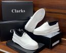Clarks men's white leather slip-on sneakers with black elastic side panels and thick white rubber sole, displayed on black branded shoeboxes.