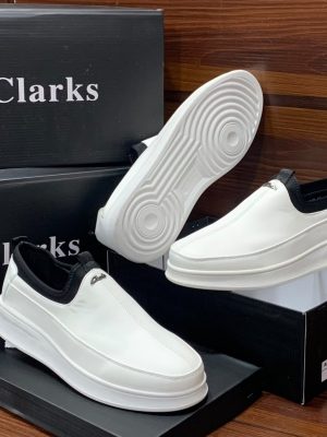 Clarks men's white leather slip-on sneakers with black elastic side panels and thick white rubber sole, displayed on black branded shoeboxes.
