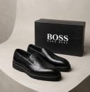 Hugo Boss men's brown leather slip-on loafers with embossed script detailing and black rubber lug sole, displayed with branded black shoebox and perfume bottle on white pedestal.
