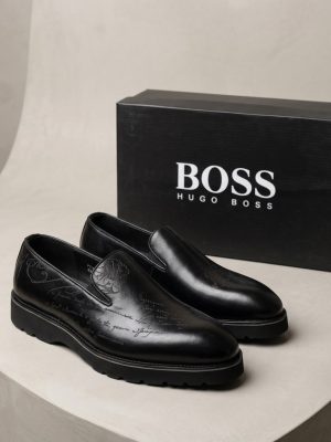 Hugo Boss men's brown leather slip-on loafers with embossed script detailing and black rubber lug sole, displayed with branded black shoebox and perfume bottle on white pedestal.