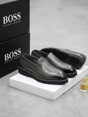 Hugo Boss men's brown leather slip-on loafers with embossed script detailing and black rubber lug sole, displayed with branded black shoebox and perfume bottle on white pedestal.