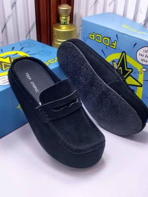 FDCP men's taupe suede penny loafer mules with braided strap detail and thick rubber sole, displayed on metallic surface against stone wall background.