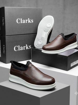 Clarks men's brown leather slip-on sneakers with elastic side panels and white rubber sole, displayed on grey pedestals with branded black shoeboxes.