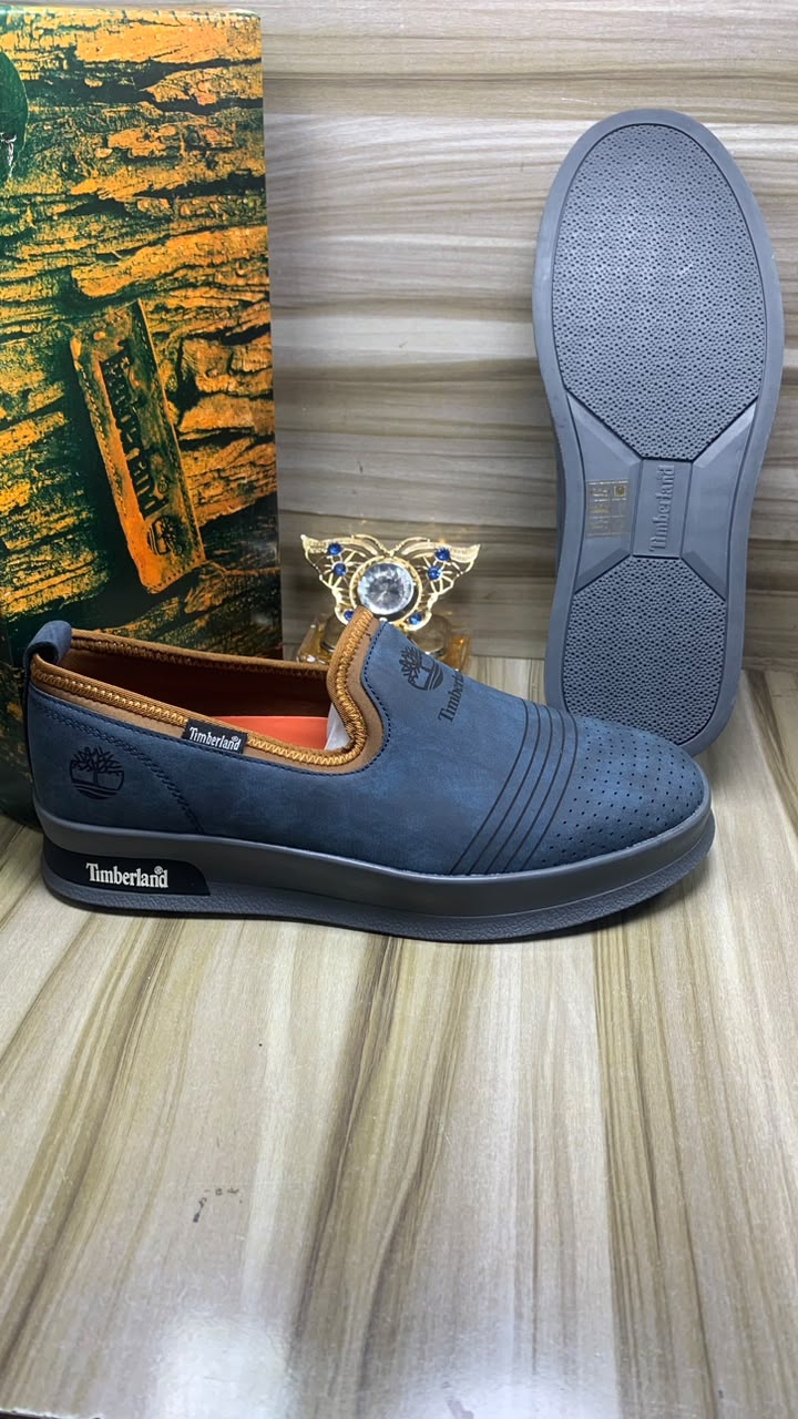 Timberland suede slip-on sneakers Timberland men's beige suede slip-on sneakers with perforated upper, navy elastic panels, and cream rubber sole, displayed on wooden floor with branded packaging.