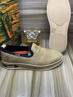 Timberland men's beige suede slip-on sneakers with perforated upper, navy elastic panels, and cream rubber sole, displayed on wooden floor with branded packaging.