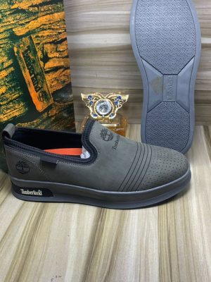 Timberland men's beige suede slip-on sneakers with perforated upper, navy elastic panels, and cream rubber sole, displayed on wooden floor with branded packaging.