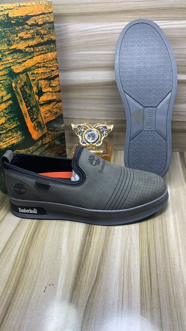 Timberland suede slip-on sneakers Timberland men's beige suede slip-on sneakers with perforated upper, navy elastic panels, and cream rubber sole, displayed on wooden floor with branded packaging.