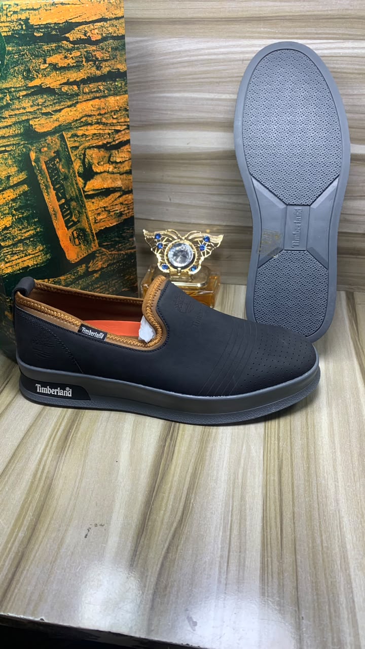 Timberland beige suede slip-on sneakers Timberland men's beige suede slip-on sneakers with perforated upper, navy elastic panels, and cream rubber sole, displayed on wooden floor with branded packaging.